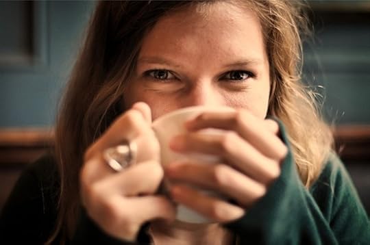 A young woman holds a coffee cup in front of her mouth. You can tell from her eyes that she's smiling.