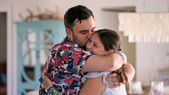 A man wearing a colorful floral shirt lovingly hugs and kisses a woman on the cheek inside a cozy, well-lit home. The woman smiles warmly with her eyes closed, and a chandelier and pastel furniture can be seen in the background.