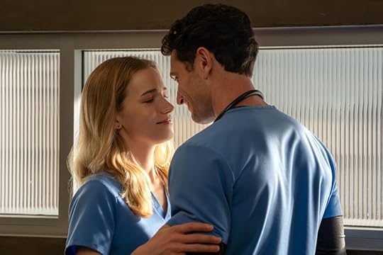 A man and woman wearing blue medical scrubs stand close together in front of a window with frosted glass, sharing an intimate and affectionate moment as they smile and gaze into each other’s eyes. A stethoscope hangs around the man’s neck.