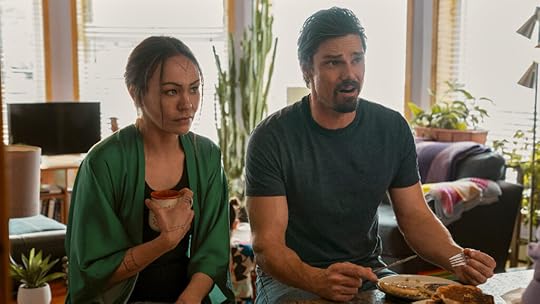 A woman in a green robe with holds a mug while sitting next to a man in a dark t-shirt, who gestures expressively with a fork in hand as they sit at a breakfast counter. Plates of pancakes are in front of them, and the cozy living room behind them is filled with sunlight, houseplants, and colorful textiles, giving the space a warm and lived-in feel.