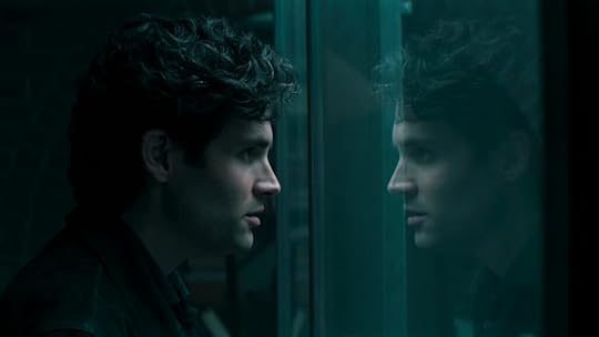 A man with dark, curly hair stares intently at his own reflection through a glass wall in a dimly lit, teal-toned space. The mood is tense and introspective, highlighting an internal struggle or moment of deep contemplation