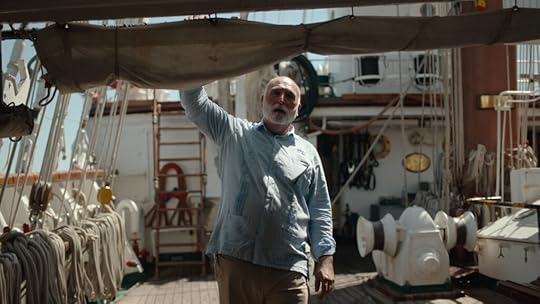 A man with a gray beard and light blue shirt stands on the deck of a ship, holding onto a rolled-up sail above him. He is surrounded by ropes, nautical equipment, and wooden features of the boat, bathed in daylight.