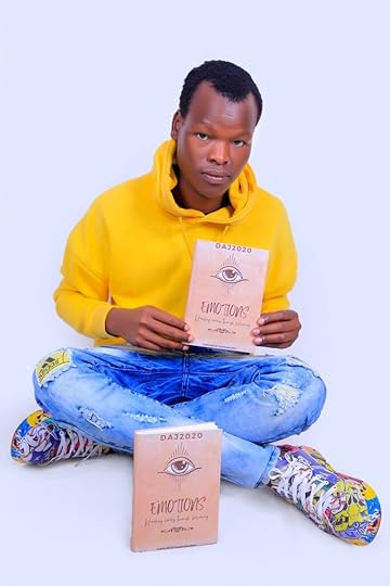 The author, a Black man with short hair, clean shaven, with a calm and neutral expression, in a yellow hoodie, distressed effect blue jeans and colourful paint splatter effect trainers, sitting cross-legged on the floor against a plain background holding a copy of EMOTIONS and with another copy upright on the floor in front of him.