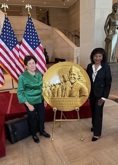 Two women stand on either side of a large gold medallion honoring the Women’s Army Corps, featuring engraved images of WWII servicewomen. Two American flags are displayed behind them. A statue is partially visible on the right in a spacious, formal setting.