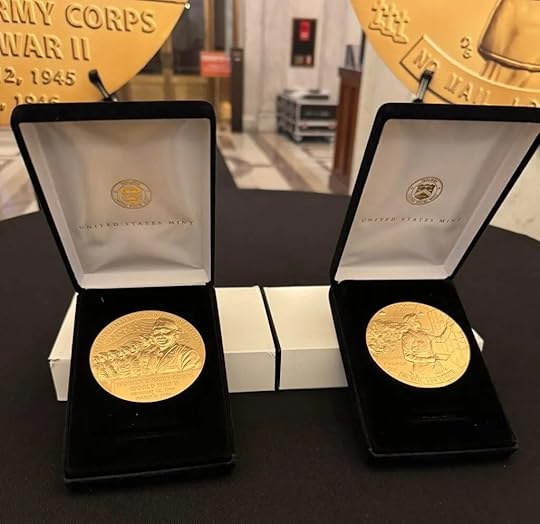 Two gold-colored commemorative medals in black velvet display boxes sit on a table. Each medal is engraved with images and text, with 'United States Mint' printed inside the box lids. In the blurred background are large medallions and a marble hallway.