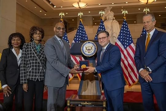 Five people in formal attire stand in a grand hall with four U.S. flags and a large statue in the background. Two men in the center hold a medal together in front of a podium displaying the U.S. House of Representatives seal.