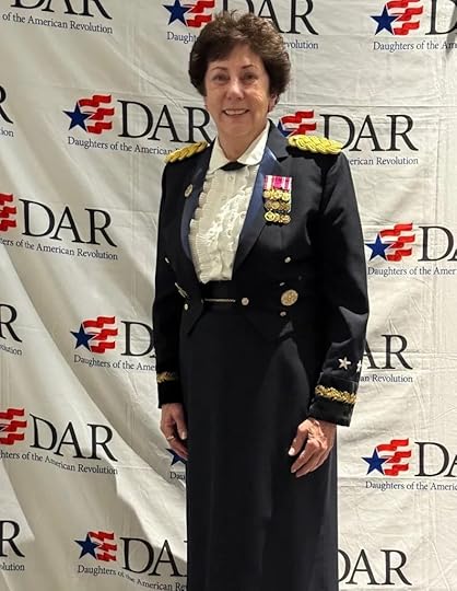 A woman in a formal military-style uniform with medals stands, smiling, in front of a backdrop featuring repeating 'DAR' logos and the words 'Daughters of the American Revolution' in blue. Her uniform is navy with gold accents and she wears a white blouse.