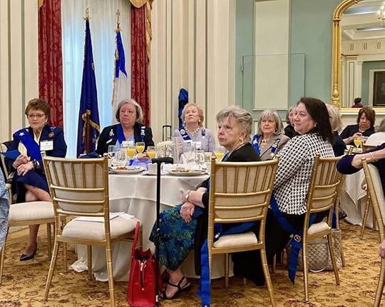A group of older women in formal attire are seated around a round table in an elegant dining room with cream-colored walls and tall red curtains. Most of the women wear blue sashes and name tags. They are listening intently, some with hands folded or resting on the table. The table is set with glasses, plates, and various breakfast foods.