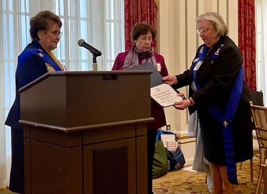 Ret. Gen. Mari K. Eder stands by a podium between two women while receiving a certificate in a formal room. Two of the women wear blue sashes; one of them is speaking at the podium, while the other is handing a certificate to a third woman who is wearing a red jacket. All three appear focused and engaged in what seems to be a ceremonial or award presentation.
