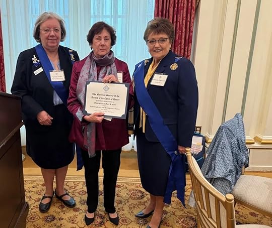 Ret. Gen. Mari K. Eder stands between two other women, all wearing dark formal clothing and blue sashes. The woman in the center holds a certificate. They stand on a patterned carpet, with a wooden chair to the right and ornate curtains and walls in the background.