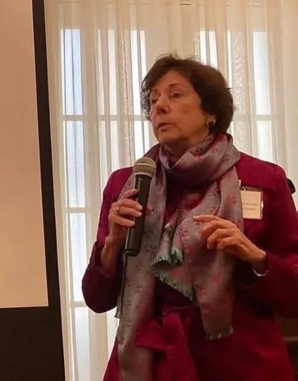 Ret. Gen. Mari K. Eder, an older woman with short brown hair is speaking while holding a microphone. She is wearing a magenta coat and a light purple patterned scarf. She stands indoors in front of sheer white curtains and a window, with part of a projection screen visible to her left.