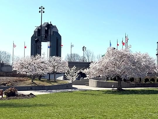 A wide lawn in a park bordered by blossoming cherry trees. In the background stands a tall, dark monument featuring a bell, with several flagpoles displaying various national flags nearby. The sky is clear and blue. Concrete walkways wind through the park.