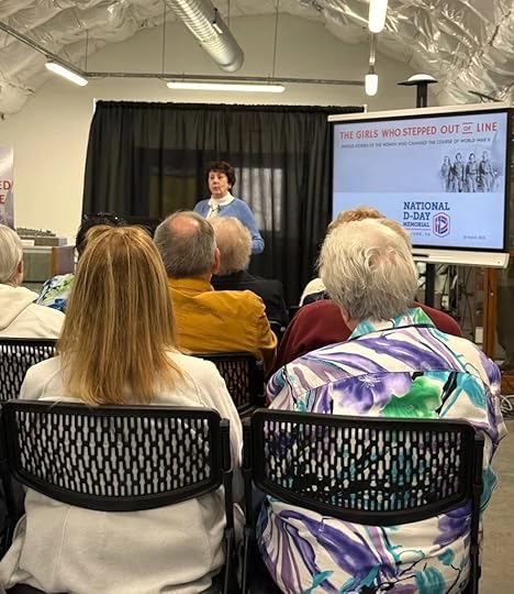 A woman stands speaking to an audience in a room with black curtains. Beside her is a poster that reads, ‘The Girls Who Stepped Out of Line: True Stories of the Women Who Changed the Course of World War II.’ Several people are seated and listening to her presentation.