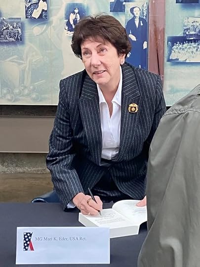A woman with short dark hair, wearing a pinstripe suit, sits at a table and signs a book. A nameplate on the table reads 'MG Mari K. Eder, USA Ret.' She is indoors, in front of a wall displaying historical photographs, and she smiles slightly while writing.