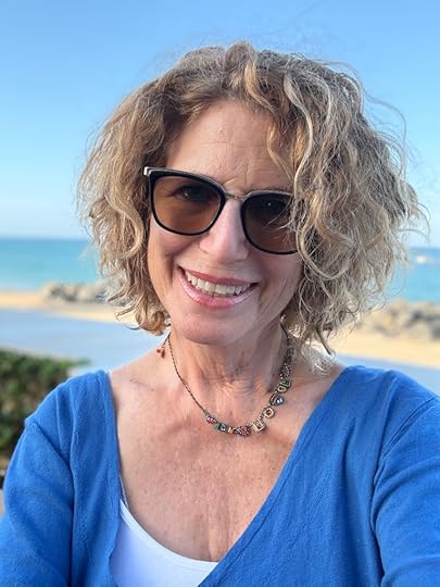 A smiling woman with curly hair wearing sunglasses and a blue top, poses for a selfie against a beach backdrop.