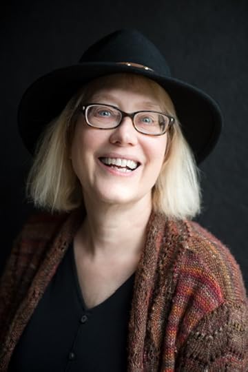A smiling woman with blond hair wearing glasses and a black hat, dressed in a colorful knitted sweater, is positioned against a dark background.