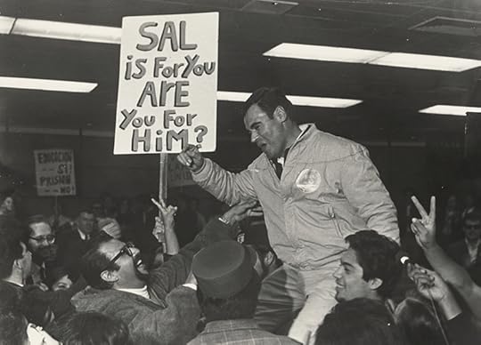 In a crowded room, people hold a smiling man wering a jacket aloft. A sign behind him reads, 