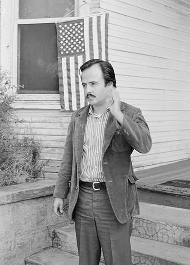 Ruiz, in dress slacks and jacket, waves to someone off camera, while standing in front of a house with an American flag hung outside a window. 