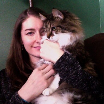 white woman with dark straight hair, wearing black and a half-smile, face half-hidden by a gorgeous fluffy cat with a tabby head and white front.