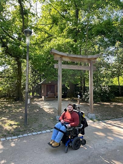 Jongeman in een rolstoel voor een Japanse poort in een park, omringd door bomen en natuurlijke omgeving.