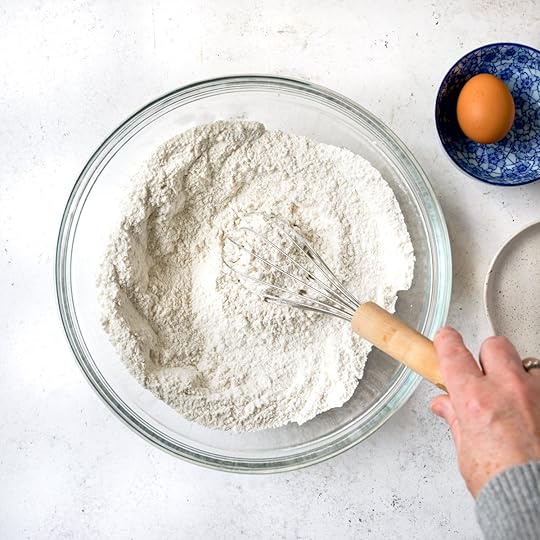 A hand holding a whisk stirring some flour in a clear bowl.