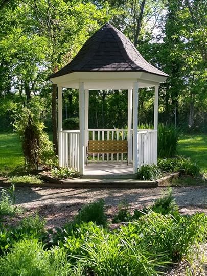 A quaint gazebo with a shingled roof and wooden bench, surrounded by lush greenery and garden plants.