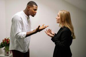 tall dark male arguing with a shorter blonde woman, his hand are out pointed palm up