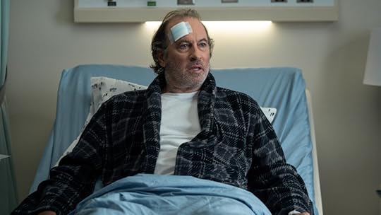 A middle-aged man with graying hair and a beard sits upright in a hospital bed, wearing a black plaid robe over a white shirt. A large bandage is taped to his forehead, and his expression is dazed and concerned as he looks off to the side. The clinical setting and medical equipment in the background emphasize the seriousness of the moment.