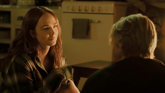 A young woman with long auburn hair sits cross-legged on the floor, wearing a green flannel shirt, and smiles warmly at a man with light blond hair, whose back is to the camera. The scene is softly lit with a golden glow, evoking a quiet, intimate fireside moment.