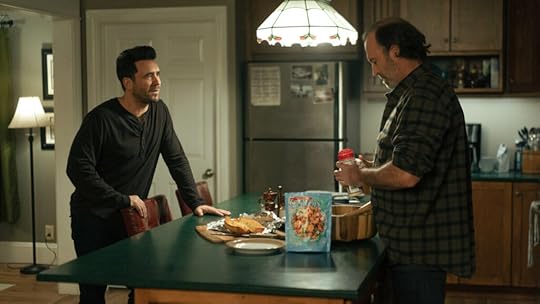 Two men talk in a warmly lit kitchen. One, dressed in a dark long-sleeve shirt, leans against a counter with a serious expression, while the other, in a plaid shirt, prepares food beside a salad bag and garlic bread.