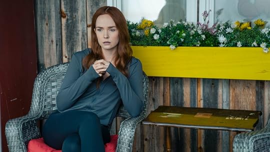 A red-haired woman sits pensively on a wicker chair outside a rustic wooden building, her hands clasped in front of her as she stares off thoughtfully. She wears a dark zip-up top and leggings, with a bright yellow flower box full of blooms behind her.