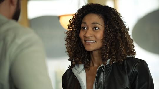 A woman with curly hair smiles warmly while speaking to someone whose back is to the camera. She wears a black leather jacket over a zip-up hoodie and stands in a softly lit café or restaurant, with round pendant lighting and blurred signage in the background.
