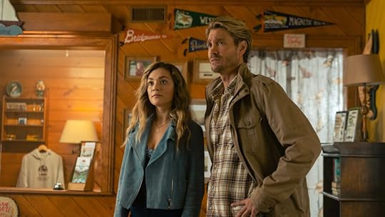 A woman with wavy blonde hair in a teal suede jacket stands beside a man with tousled blonde hair in a tan jacket and plaid shirt. They appear alert and concerned as they look off-camera inside a cozy, wood-paneled shop or lodge, decorated with pennants and vintage memorabilia.