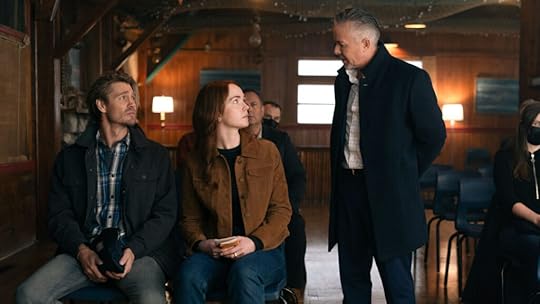 A man and woman sit side by side in a wooden hall, looking up at another man in a suit who stands beside them speaking. The seated man wears a plaid shirt and dark jacket, while the woman has red hair and wears a brown suede coat, holding a notepad in her lap. Other people sit in rows behind them at an auction.