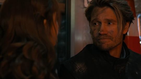 A man with dirt and soot on his face, wearing a black jacket, looks emotionally at a woman whose face is mostly out of frame. His expression is somber and reflective, suggesting the aftermath of a traumatic event. The warm lighting and background hint at an emergency scene.