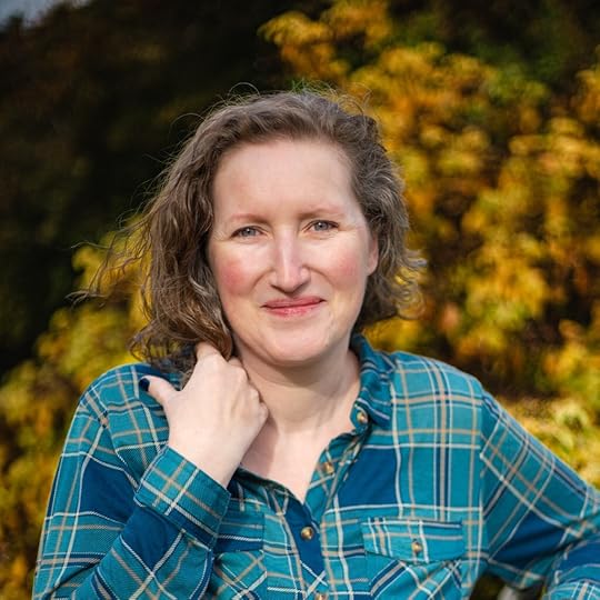 White woman in blue checked shirt smiling, with short jaw-length light brown wavy hair, and blue eyes. Blurred trees are in the background.