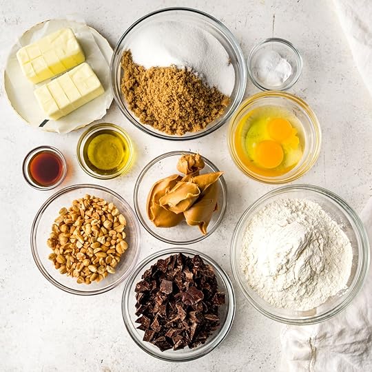 All of the ingredients you need to make peanut butter chocolate chip cookies in bowls on a white surface.