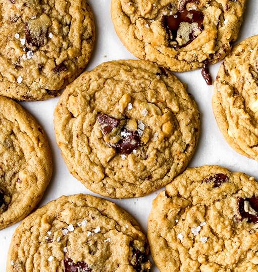 Close up overhead photo of peanut butter cookies with chunks of glossy chocolate, peanuts and sea salt flakes on a white surface.