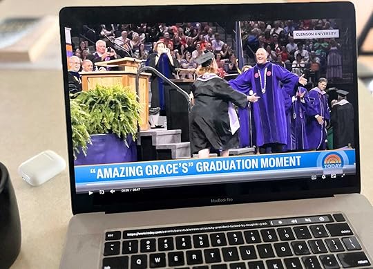  a laptop screen displays a graduation ceremony with a graduate in a cap and gown hugging an older man in academic regalia, with the text “Clemson University” and “GRACING GRACE'S GRADUATION MOMENT” on the screen. 