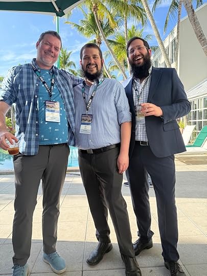 Three men are standing together outdoors, smiling at the camera. They are wearing name tags and appear to be at an event. One man is in a plaid shirt, another in a striped shirt, and the third in a suit. They are holding drinks, and there are palm trees and a pool in the background.