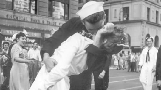 A black-and-white photograph depicting a sailor dressed in a traditional naval uniform passionately kissing a woman in a white dress on a busy city street. The scene is lively, with pedestrians and other sailors visible in the background, suggesting a celebratory atmosphere. Buildings, including the Chemical Bank & Trust Co. and Walgreens Drug Store, are seen in the background, adding to the urban setting.