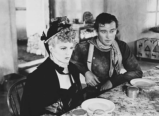 Claire Trevor in a black hat and dress and John Wayne in what looks like a Union frock coat sit at a dining table in