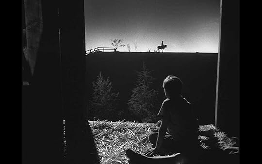 A child watches a man on a horse on the horizon from a bale of hay in a barn in