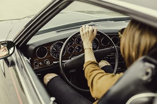 A woman in a gray car with her hand on the steerring weehl, a gold bracelet on her wrist.
