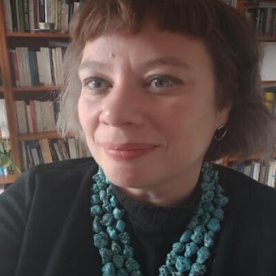 Close-up portrait of a woman with short brown hair, wearing a black top and a large turquoise necklace, set against a background of bookshelves filled with books.