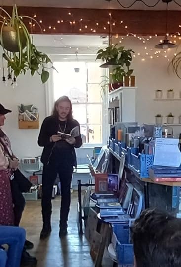 Image shows author Die Booth, a medium-built white man with long dark hair and dark facial hair, dressed all in black, standing at the end of a small bookshop, reading from a book. There are fairy lights on the high ceiling, shelves to the left and a counter to the right, You can just about see some people sitting around the cropped edges of the shot.
