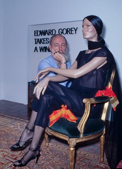 Photo shows a middle-aged man in a light blue shirt, kneeling behind an elegantly dressed mannequin, seated in a chair. He has a heavy white beard. Behind him, a sign reads 