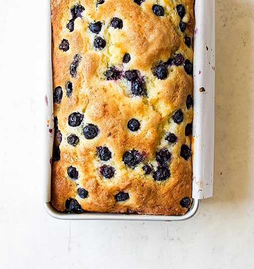A close-up, overhead shot of a golden-brown blueberry loaf, speckled with dark purple blueberries, baked in a silver loaf pan lined with white parchment paper. The loaf has a slightly domed, crinkly top.