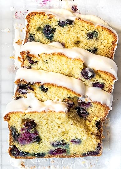 An overhead, close-up shot of a sliced blueberry lemon pound cake, drizzled with a thick white lemon glaze, arranged on a white parchment paper. The golden-yellow slices reveal a tender crumb dotted with vibrant purple blueberries and specks of lemon zest.