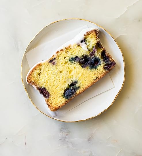 An overhead shot of a single slice of blueberry lemon loaf cake, topped with a white glaze, resting on a small white plate with a gold rim. The cake slice is golden yellow with visible dark blueberries and sits on a piece of white parchment paper on a light marble surface.